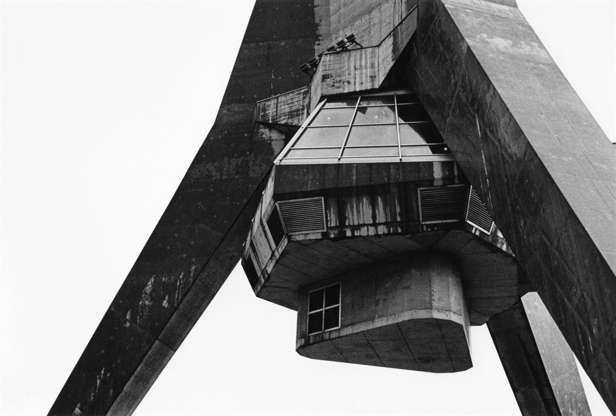 Looking up at a brutalist concrete tower and its suspended observation pod, in black and white